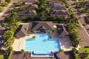 Vista aérea de um resort com uma piscina central, rodeada por vários edifícios tipo bangalô, palmeiras e jardins bem cuidados.