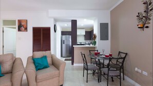 A modern living room with a sofa and chairs, a dining area with a table and chairs, and a kitchen in the background. - I Heart JA Affordable Luxury at The Bromptons (Kingston)