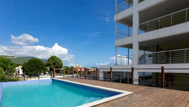 A rooftop swimming pool with a wooden deck, surrounded by a modern building with balconies. - I Heart JA Affordable Luxury at The Bromptons (Kingston)