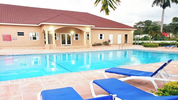 A swimming pool with blue lounge chairs, a building with a red roof, and palm trees in the background. - Serenity Retreat Villa Drax Hall (Ocho Rios)