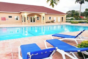 A swimming pool with blue lounge chairs, a building with a red roof, and palm trees in the background.