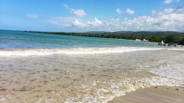 A beach with clear turquoise water, gentle waves, and a distant view of a forested coastline. - I Heart JA Fairway (Kingston)