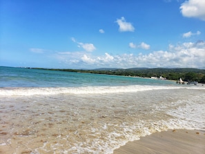 Ein Strand mit klarem türkisblauem Wasser, sanften Wellen und einer entfernten Aussicht auf eine bewaldete Küste.