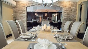 A dining area with a long table set for six, surrounded by chairs, with a stone wall and a kitchen in the background.