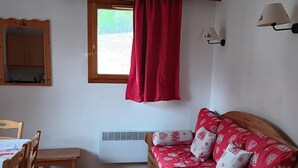 A room with a wooden sofa, a dining table with chairs, a window with a red curtain, a mirror, and wall-mounted lamps. - Wifi, 300m from ski slopes, 3rd floor, swimming pool, balcony, parking, tv, ski locker, 35m² (Puy-Saint-Vincent)
