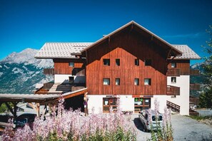 A wooden building with a ski shop, a parking area, and mountains in the background. - Wifi, 300m from ski slopes, 3rd floor, swimming pool, balcony, parking, tv, ski locker, 35m² (Puy-Saint-Vincent)