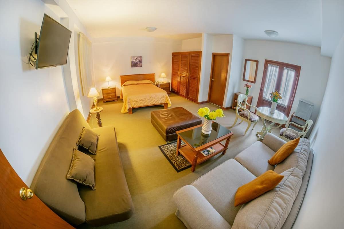 A well-lit, modern hotel room with a sofa, a bed, a dining table, and a TV mounted on the wall.