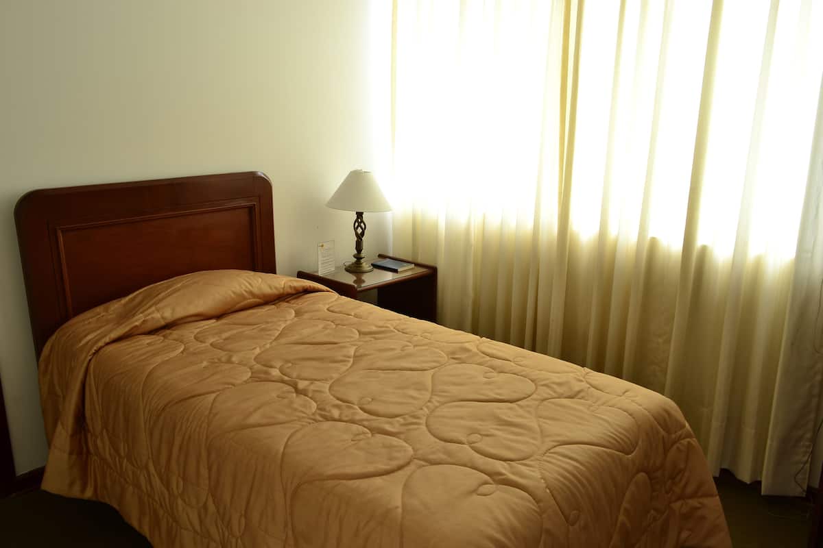 A neatly made bed with a brown quilt, a wooden headboard, a bedside lamp, and a window with sheer curtains.