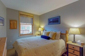 Una cama bien hecha con un almohadón azul, una lámpara de noche y un cuadro en la pared.