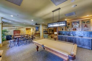 Una mesa de billar con asientos alrededor, una zona de bar con estantes llenos de objetos decorativos y un área de sala informal con chimenea.