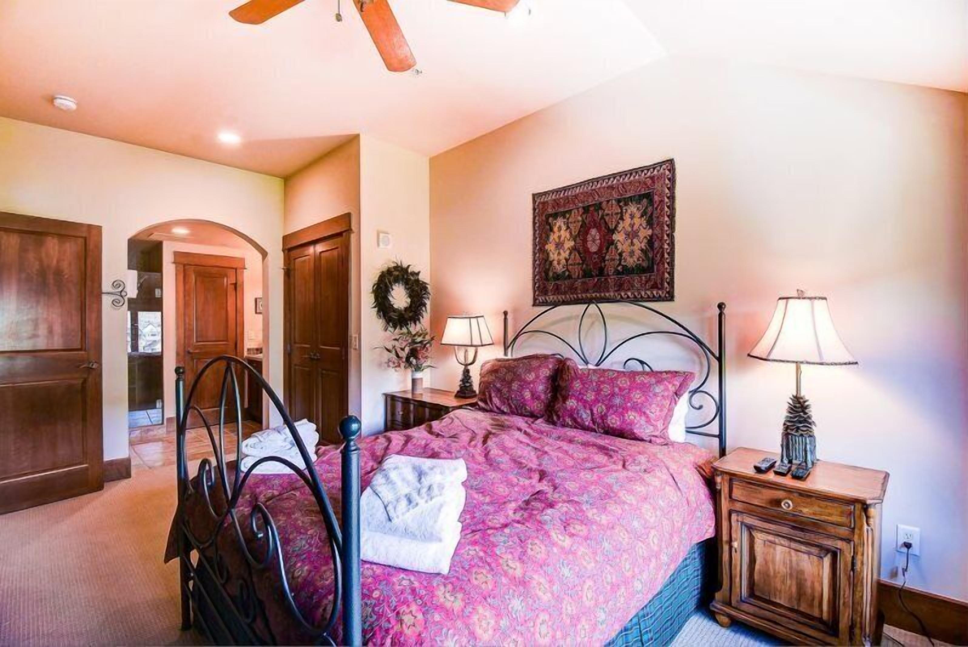A bedroom with a bed, two bedside tables with lamps, a headboard, a wall-mounted tapestry, and a door leading to another room.