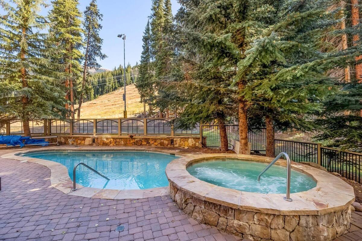 A swimming pool with a hot tub, surrounded by trees and a stone wall.