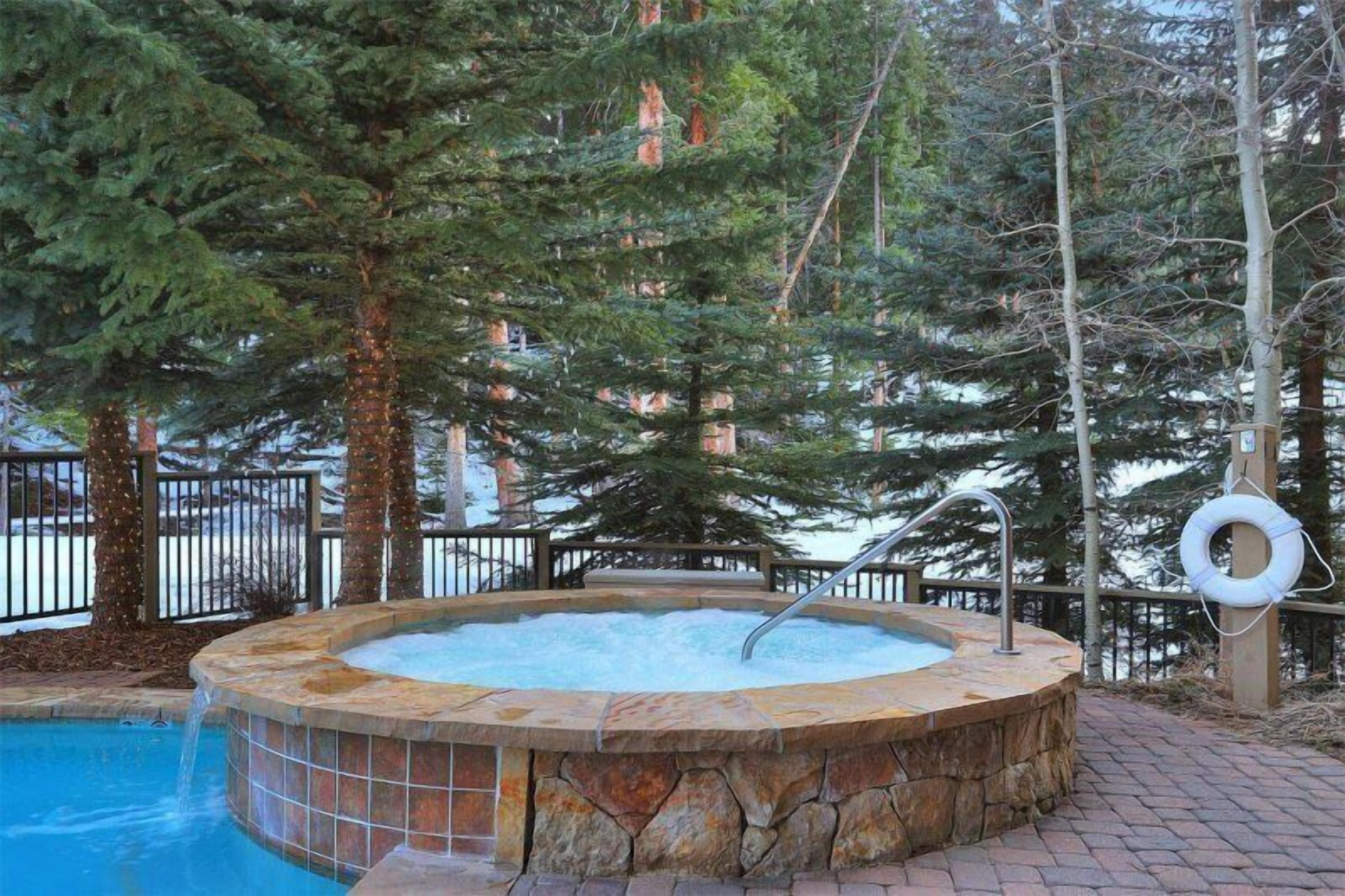 A hot tub surrounded by stone walls and a pool, with trees and a fence in the background.