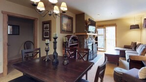 Comedor con mesa de madera, sillas y candelabro. Hay una chimenea con un televisor encima. Se ve un sofá y una mesita de noche al fondo.