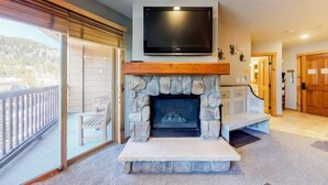 A stone fireplace with a flat-screen TV above it, a balcony with a wooden railing, and a view of a mountainous landscape. - 1 Bedroom Premier Mountain Condo in River Run Village Just a Short Walk to Shopping, Dining, and Skiing (Keystone)