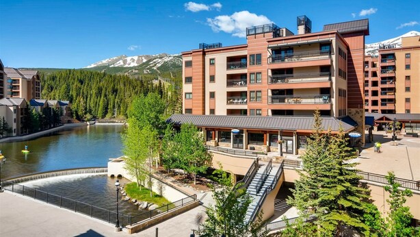 A multi-story hotel with balconies, a river view, and a mountainous backdrop.