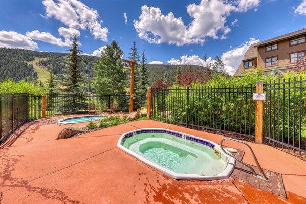A hot tub area with a pool, surrounded by a fence and trees, with mountains in the background.