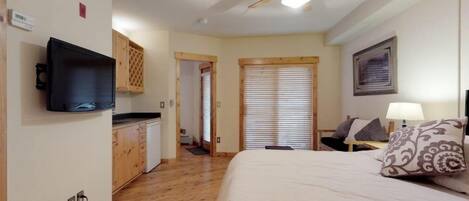 A bedroom with a bed, a ceiling fan, a TV mounted on the wall, a wooden cabinet, and a window with blinds.