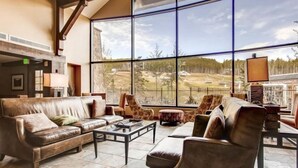 A spacious living area with leather sofas, a coffee table, and chairs. Large windows offer a view of a park and a building with a glass facade.