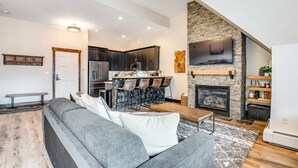 Un salon moderne avec un canapé gris, une cheminée et un coin cuisine doté de meubles sombres et d’un mur d’accent en pierre.