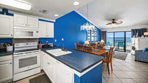 Une cuisine moderne avec des meubles blancs, un mur d’accent bleu, un coin repas avec une table et des chaises, et une vue sur l’océan grâce à de grandes fenêtres.