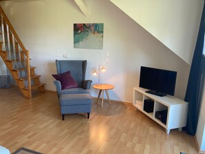 Una sala moderna con una escalera de madera, un sillón azul, una mesita redonda con una lámpara y un televisor sobre un mueble blanco.