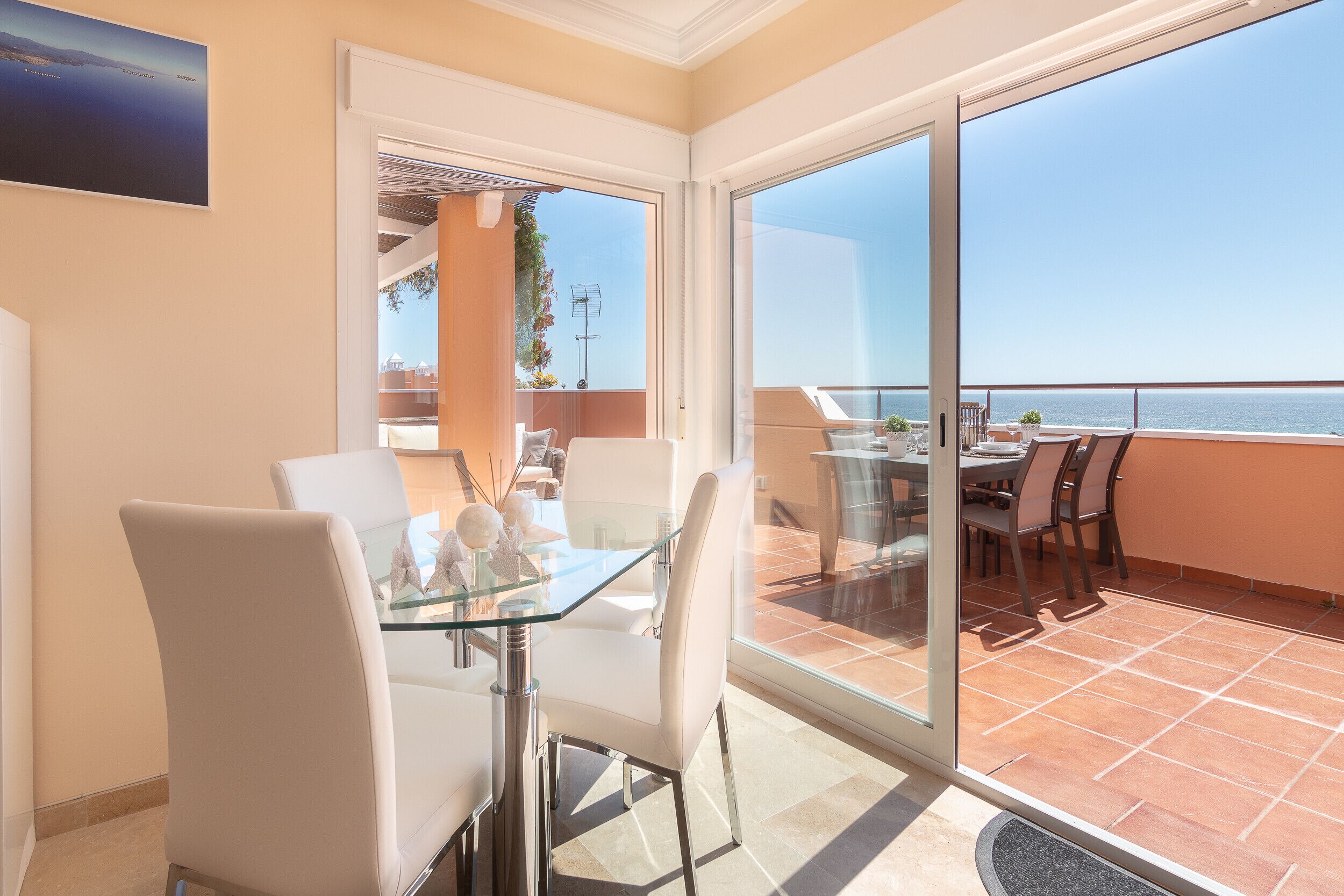 Uma zona de refeições moderna com mesa de vidro e cadeiras brancas, uma varanda com vista para o mar e uma porta que dá para outro quarto.