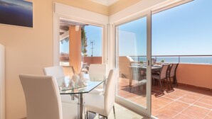 A modern dining area with a glass table and white chairs, a balcony with a view of the sea, and a door leading to another room.