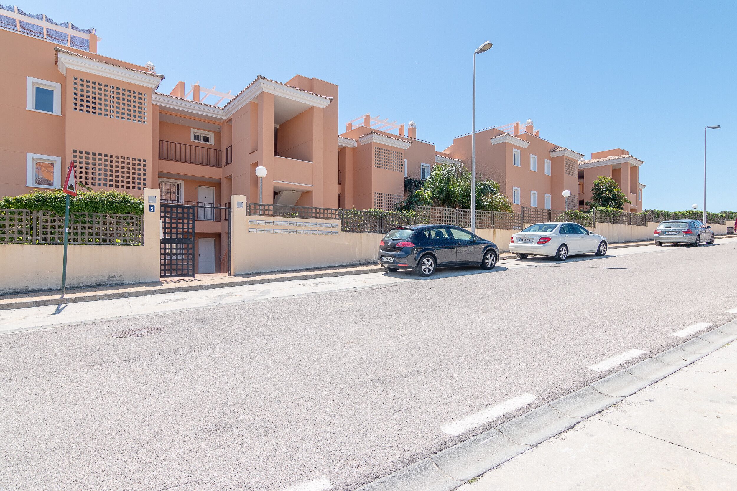 Una calle residencial con modernos edificios de apartamentos, un acceso con puerta de reja y coches estacionados.