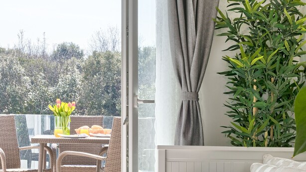 Et soveværelse med seng, et vindue med gardiner, en potteplante og en balkon med bord og stole.