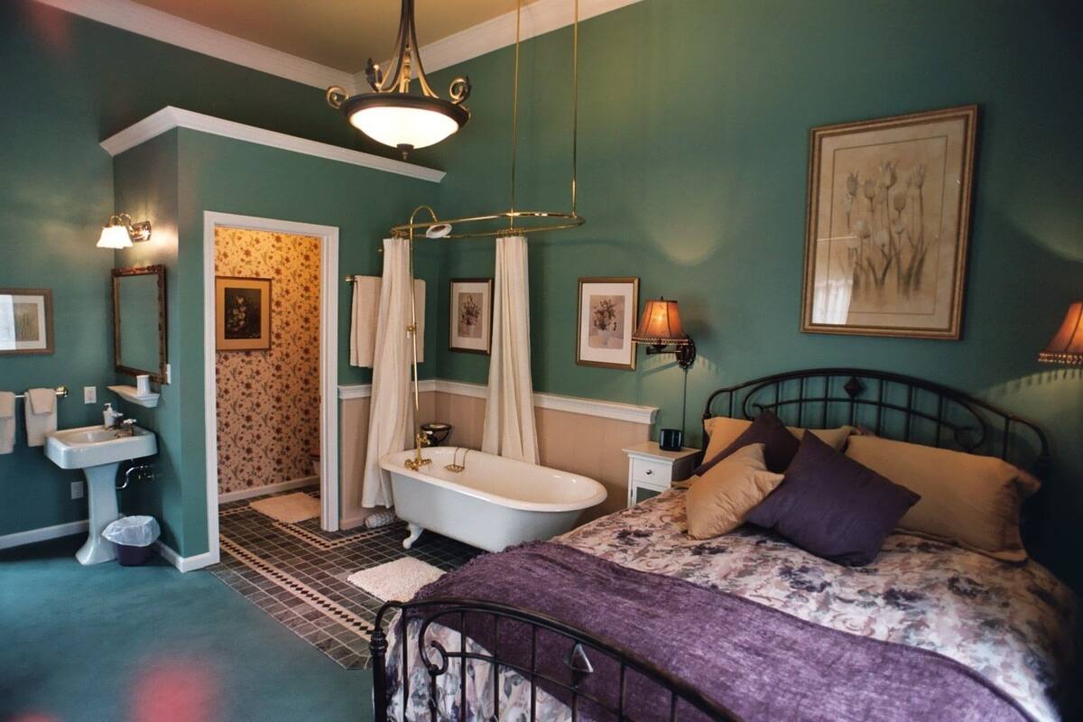 A bedroom with a bed, a nightstand, a dresser, a vanity, a bathtub, and a shower curtain.