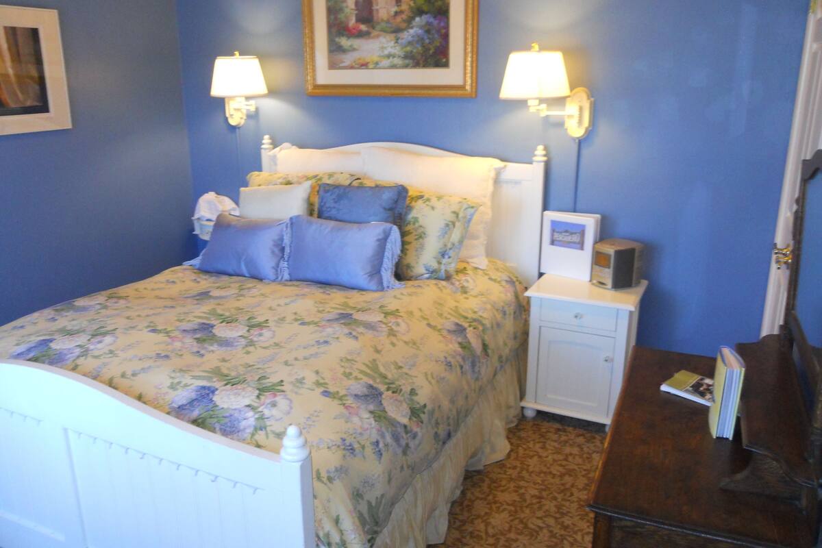 A bedroom with a floral bedspread, a white headboard, two bedside lamps, a nightstand with a lamp, and a framed picture above the bed.
