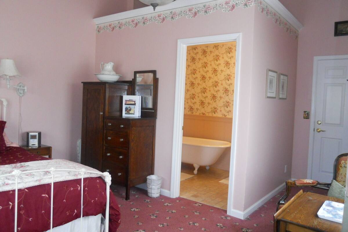 A bedroom with a bed, a dresser, a mirror, a nightstand, a lamp, and a door leading to a bathroom with a bathtub.