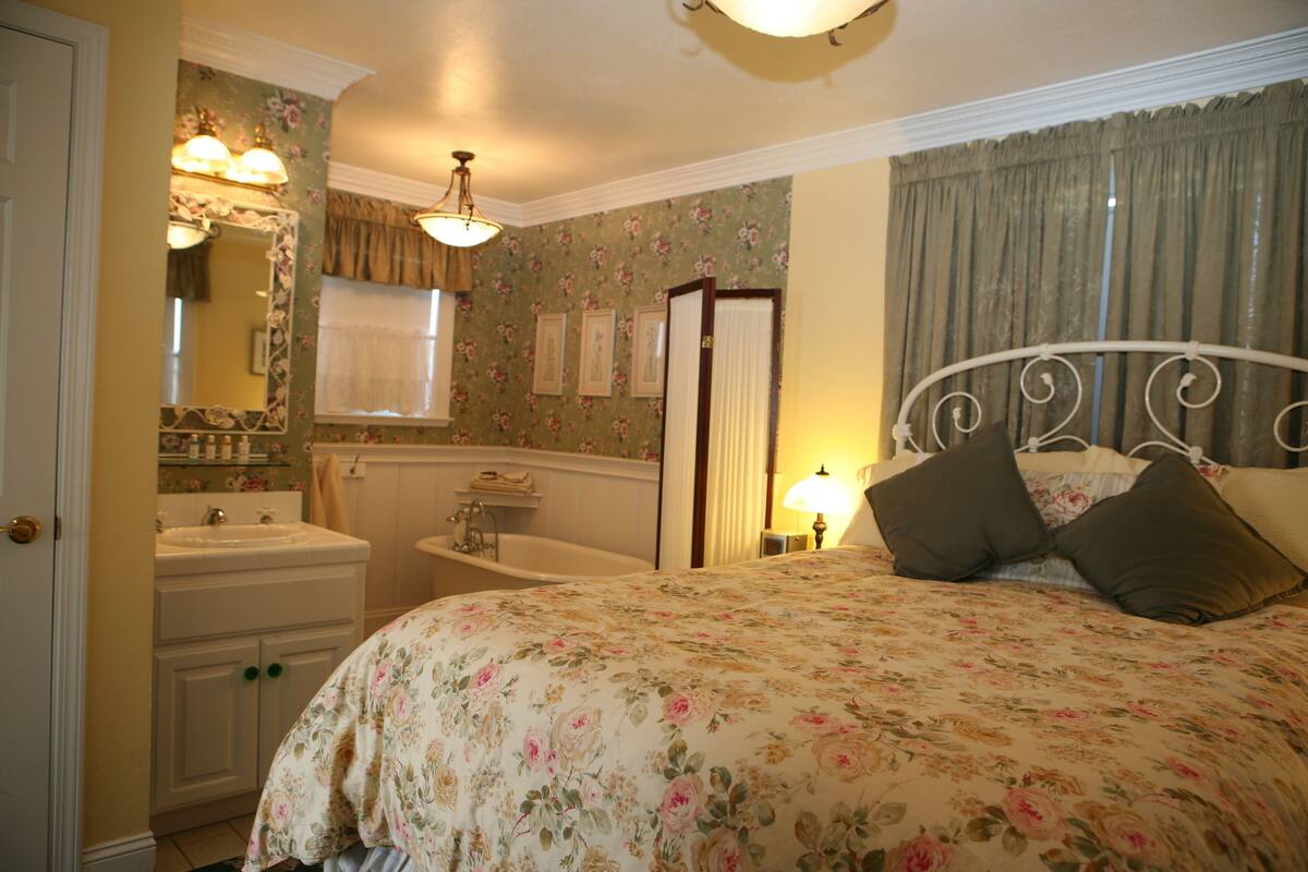 A bedroom with a floral bedspread, a white metal bed frame, a nightstand with a lamp, a bathroom with a sink and bathtub, and a mirror with floral wallpaper.