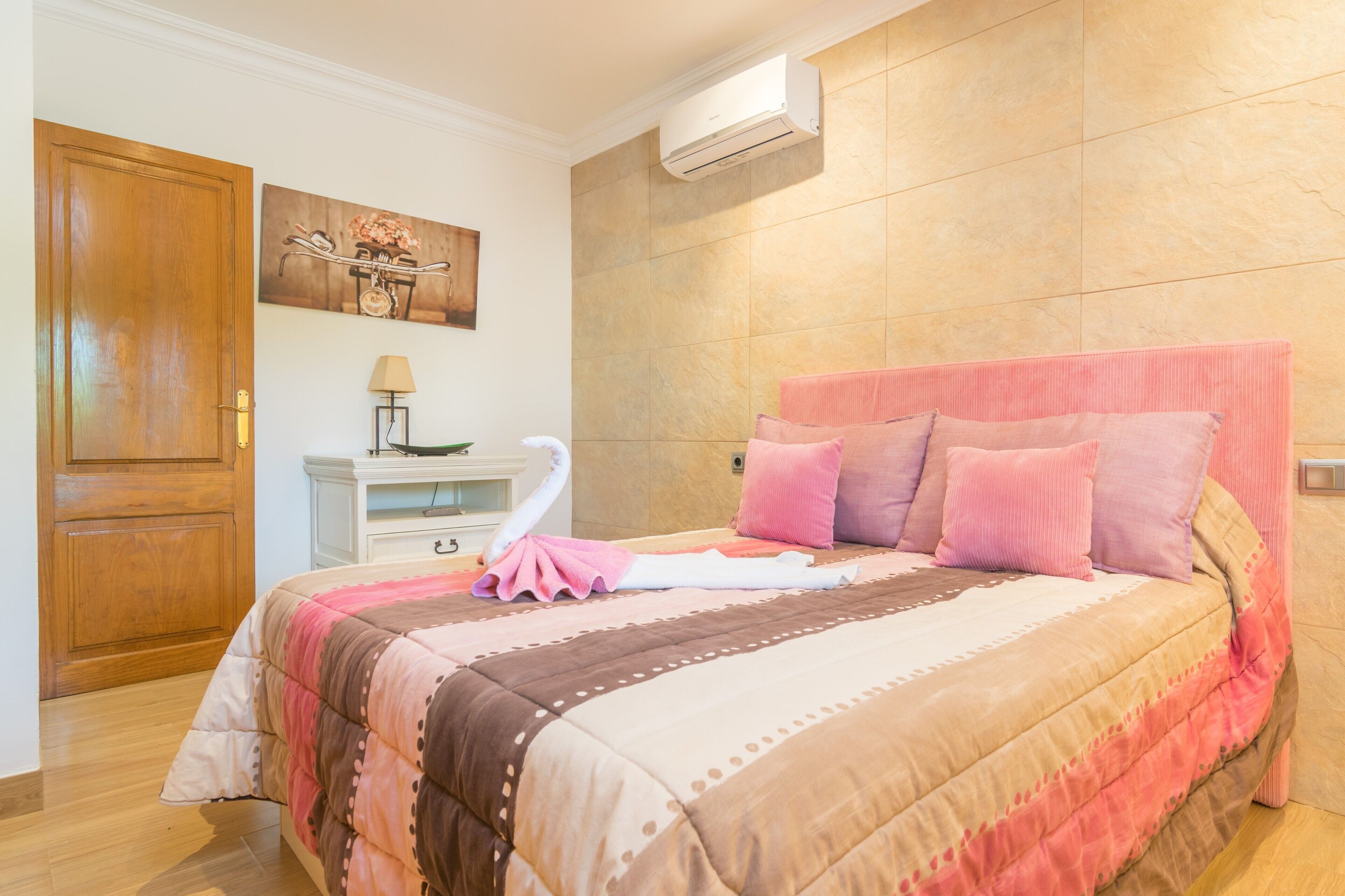 Uma cama bem arrumada com roupa de cama rosa e marrom, uma porta de madeira, um ar condicionado fixado na parede e uma pequena mesa de cabeceira com uma luminária.