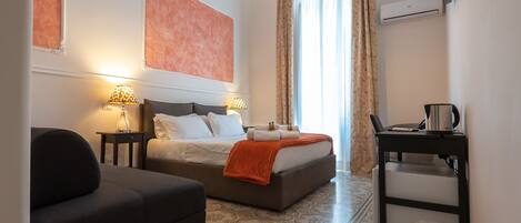 A hotel room with a bed, a sofa, a small table with a lamp, and a trash bin.