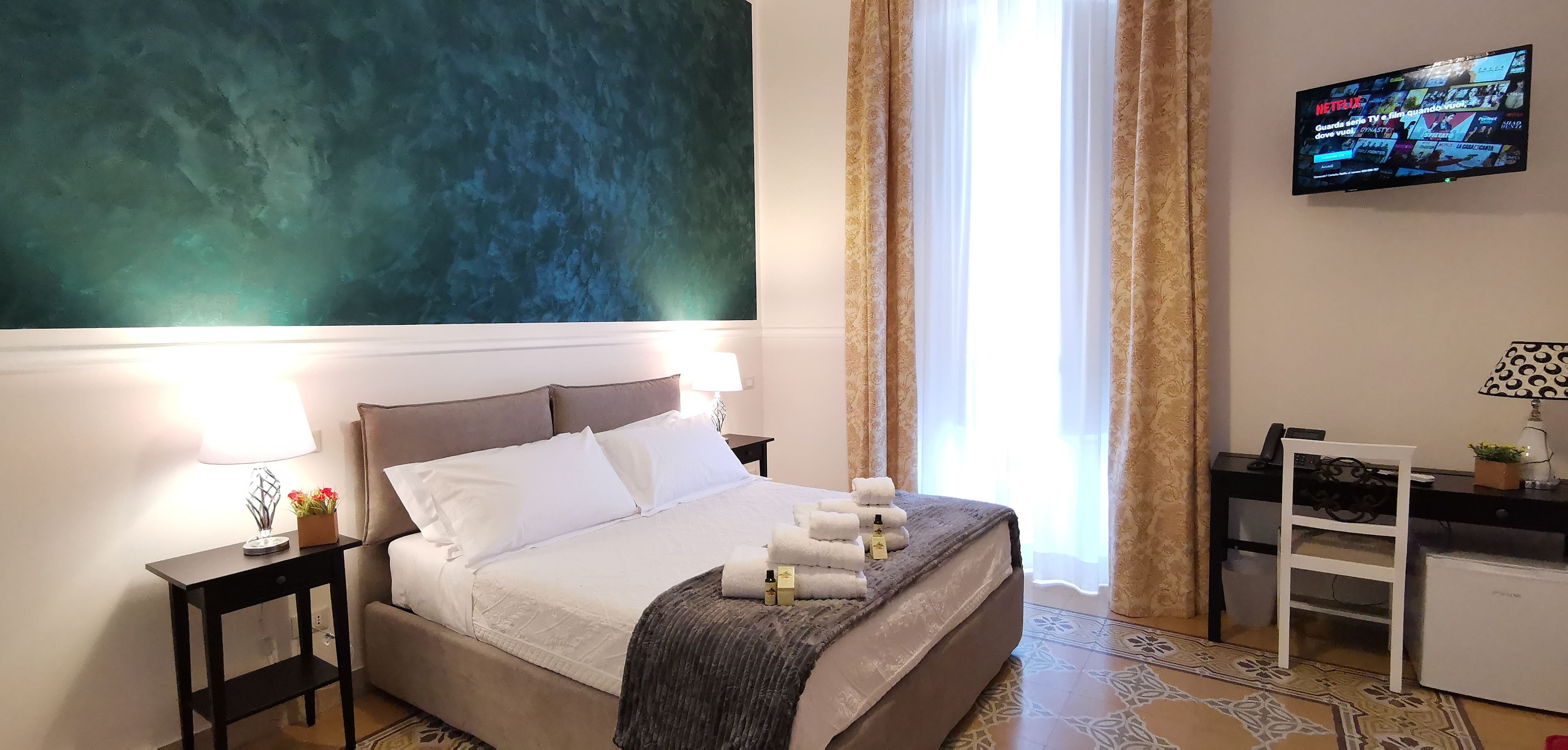 A hotel room with a bed, bedside tables, a desk, a TV, and curtains.