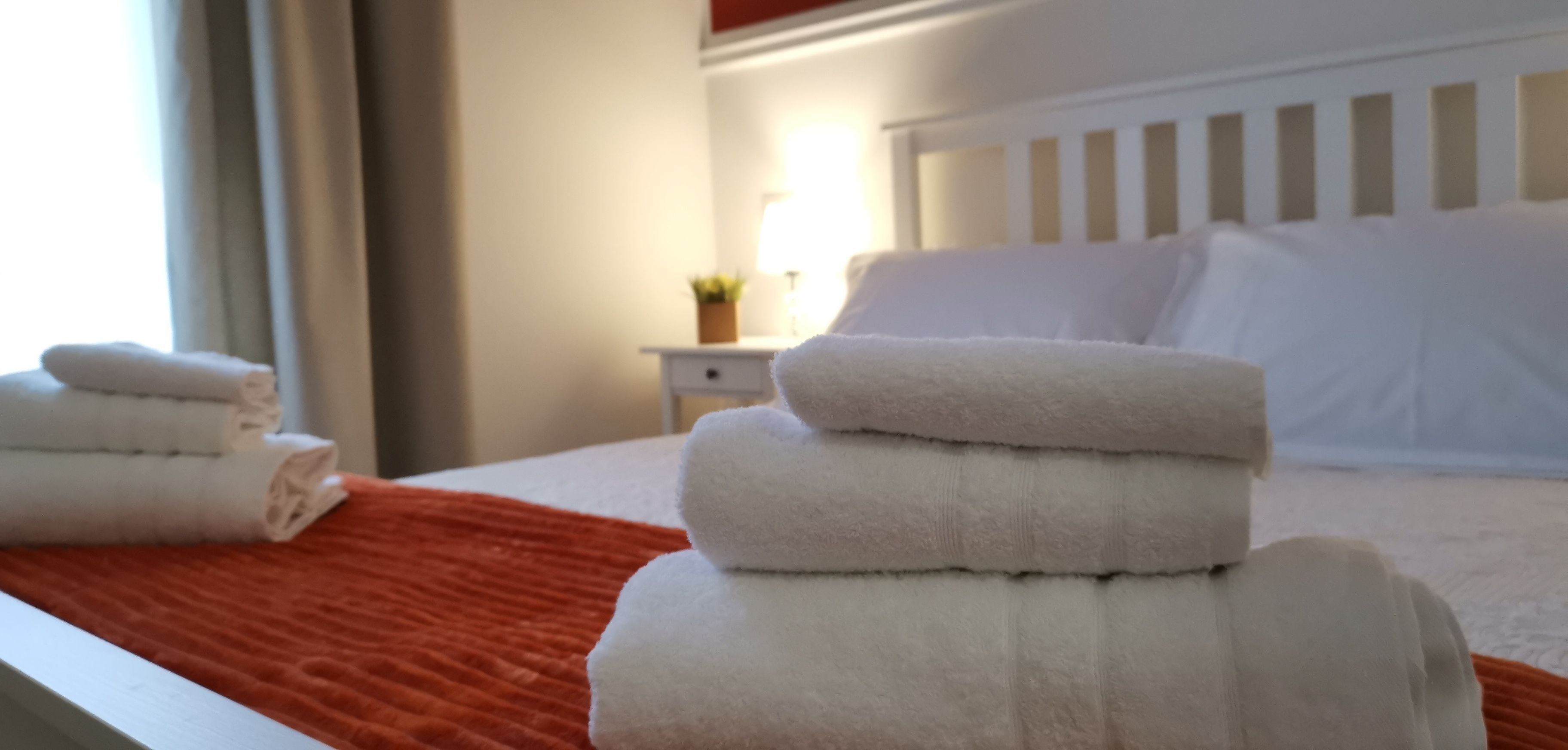 A neatly made bed with white linens and a red blanket, a small bedside table with a lamp, and a window with curtains.