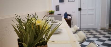 Una habitación de hotel con piso a cuadros, una mesa de comedor con un jarrón de flores amarillas y una puerta al fondo.