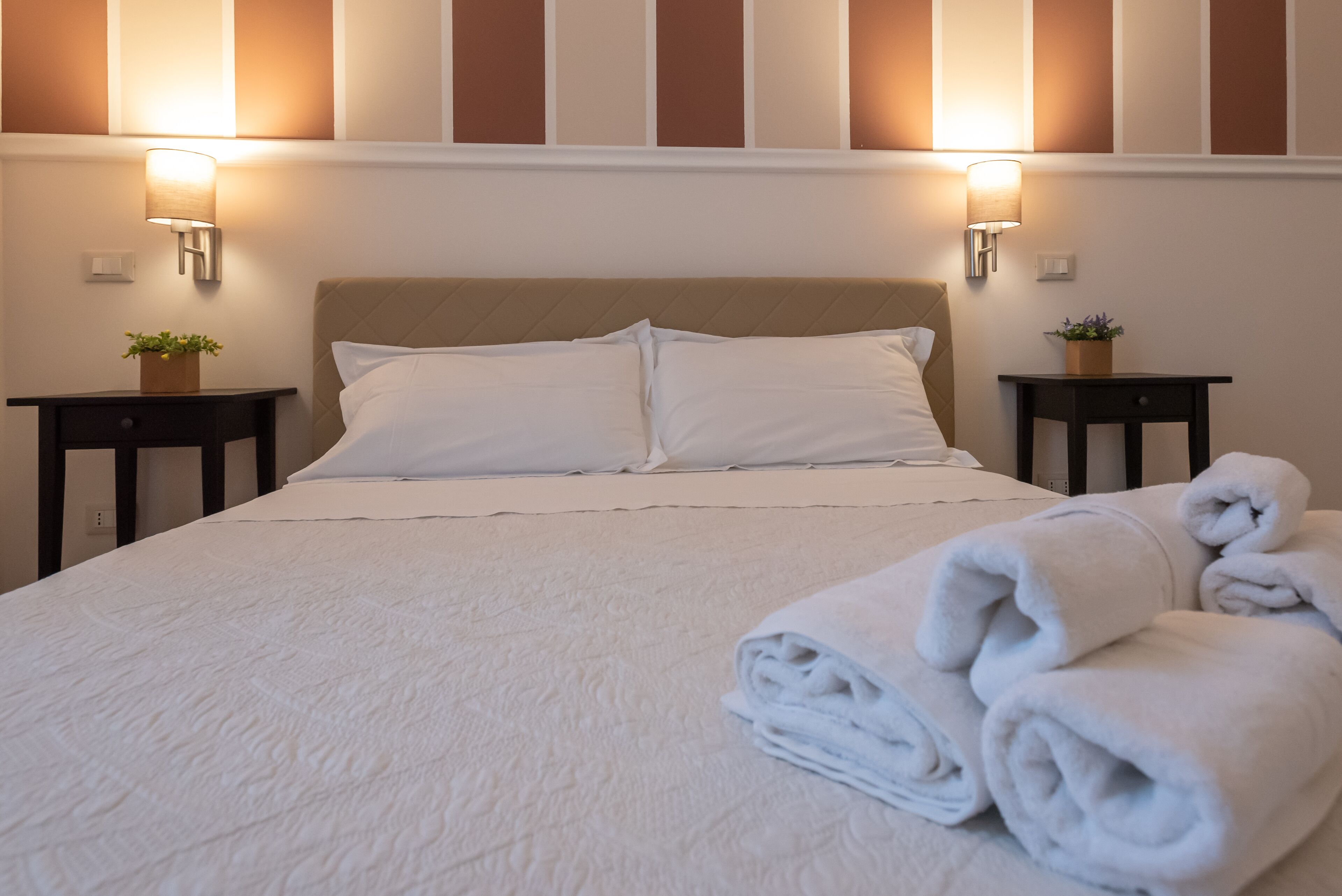 A neatly made bed with white linens, two bedside tables with lamps, and a stack of folded towels.