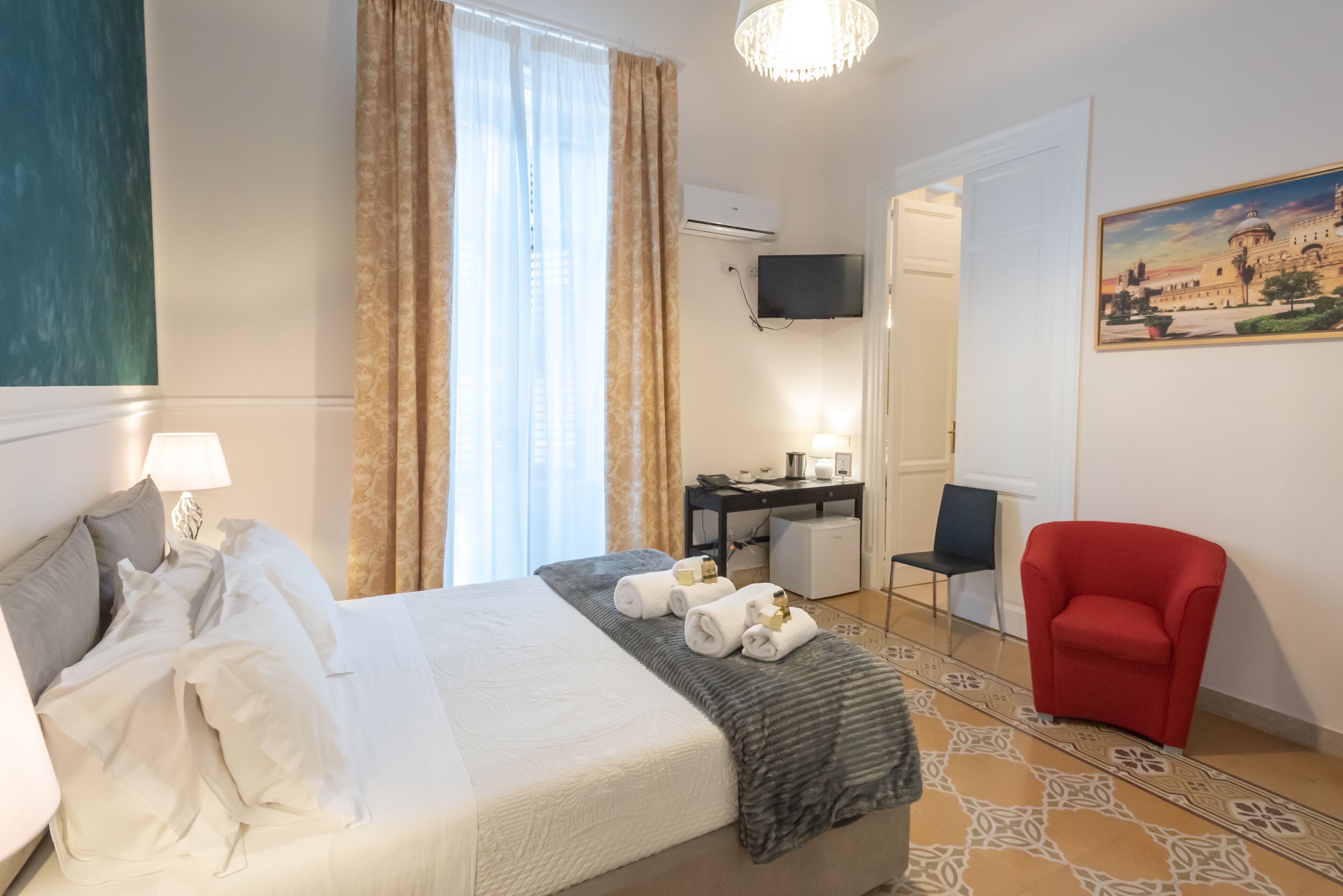 A hotel room with a bed, a desk, a chair, a red armchair, and a painting on the wall.