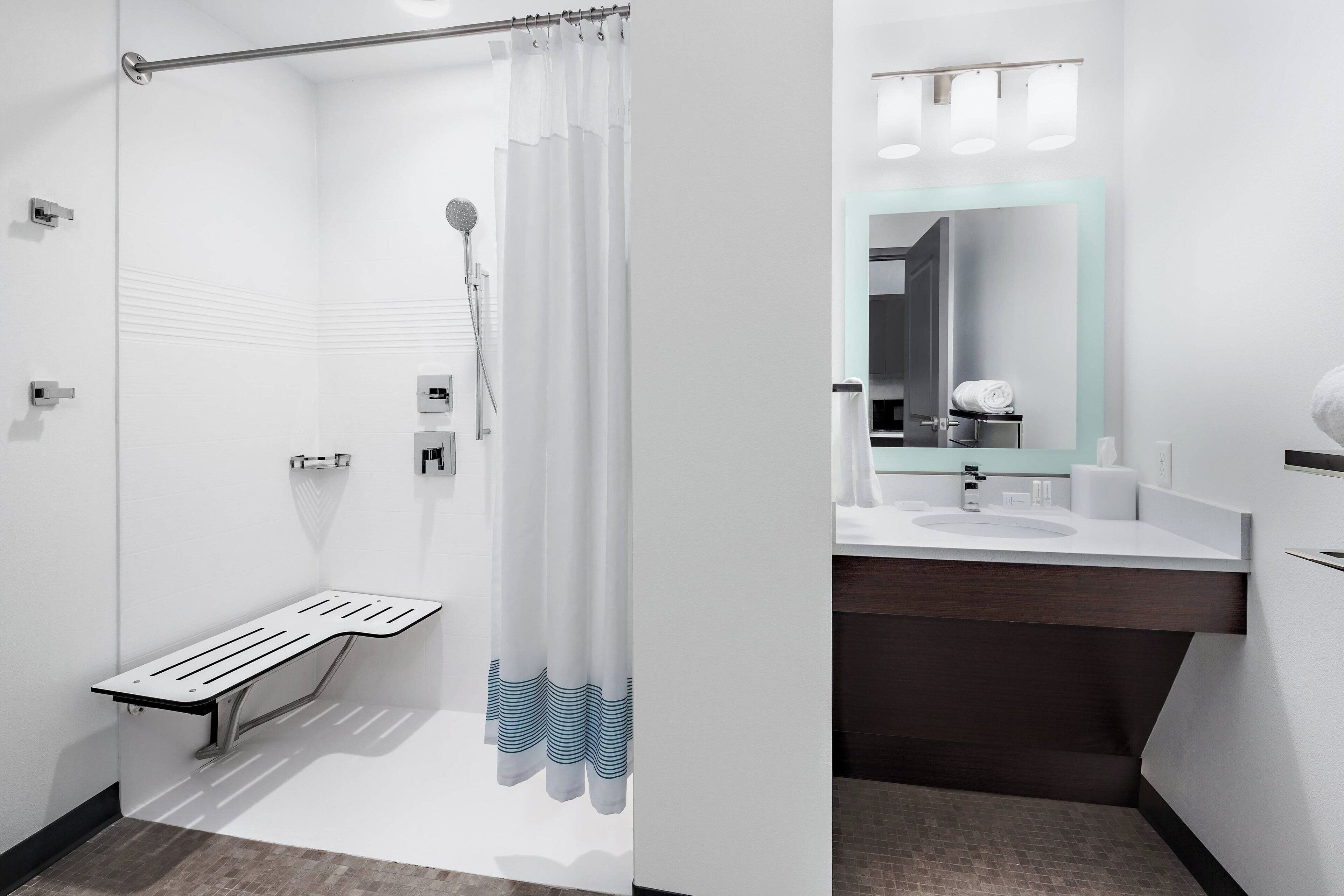 Un baño moderno con cabina de ducha de vidrio, un banco, toallero y tocador con espejo.