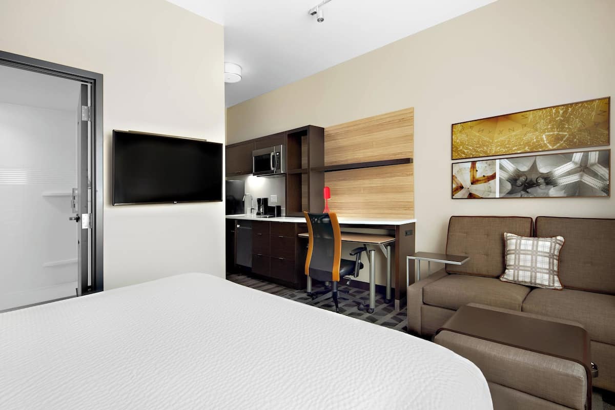 A hotel room with a bed, a desk with a computer, a kitchenette, a sofa, and a TV.