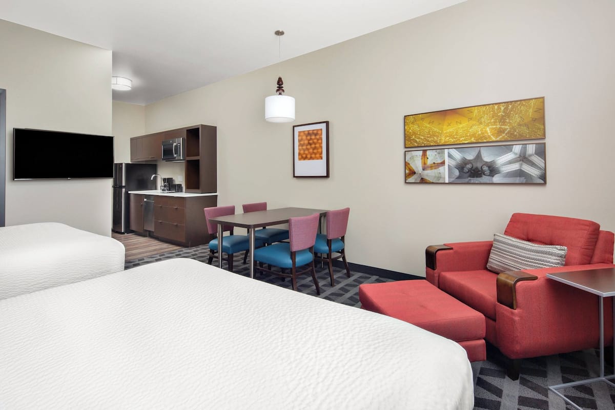 A hotel room with two beds, a dining table with chairs, a kitchenette, and artwork on the walls.