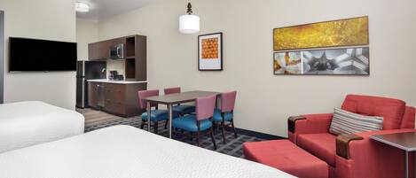 Habitación de hotel con dos camas, una mesa de comedor con sillas, una kitchenette y obras de arte en las paredes.