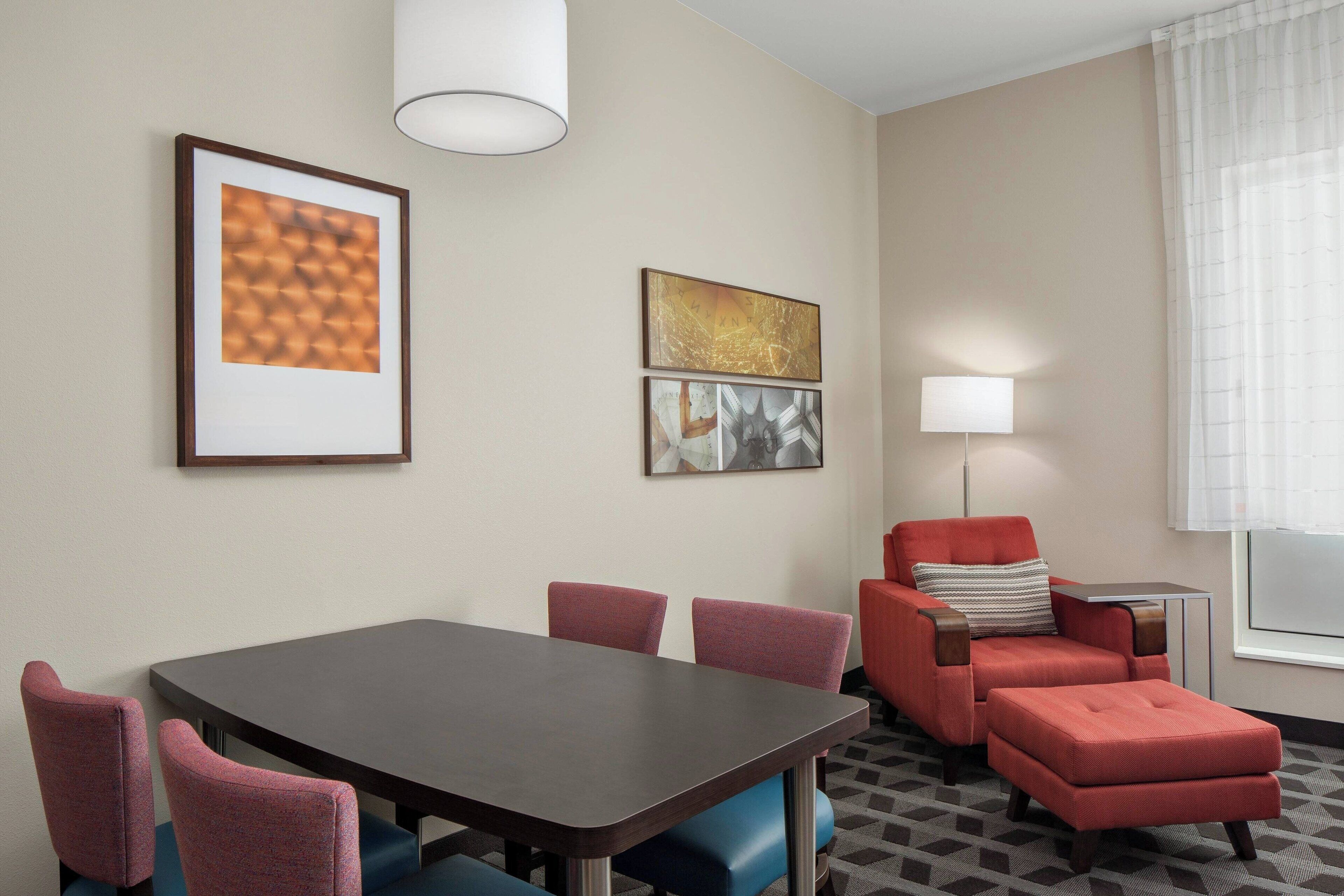 A hotel room with a dining table, red upholstered chairs, a red armchair with a pillow, a side table, and framed artwork on the wall.