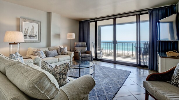 Een ruime woonkamer met een groot raam met uitzicht op een strand, ingericht met een bank, fauteuils en een salontafel.