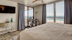 Een slaapkamer met een groot bed, een plafondventilator, een televisie aan de muur en uitzicht op het strand door het raam.
