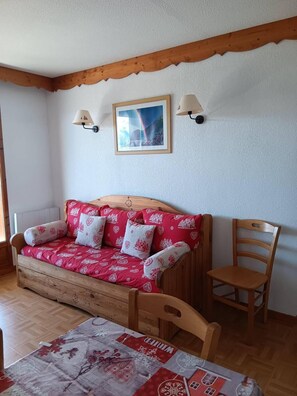 A cozy room with a wooden sofa bed, a wooden chair, a table with a patterned tablecloth, and two wall-mounted lamps. - Wifi, Ground floor, swimming pool, balcony, parking, tv, ski locker, 39m², Puy-Saint-Vincent (Puy-Saint-Vincent)