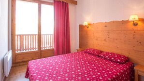 A bedroom with a wooden headboard, a bed with a red floral bedspread, two bedside lamps, and a window with a wooden balcony. - Wifi, 2nd floor, swimming pool, balcony, parking, tv, ski locker, 60m², Puy-Saint-Vincent (Puy-Saint-Vincent)